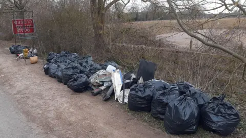 Mark Fishpool Abut 20 black bags of litter neatly stacked in a layby, with some larger items of rubbish among them.