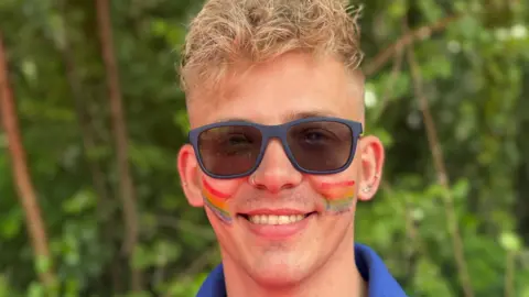 A man with blond hair is wearing grey sunglasses and a blue t-shirt. He has rainbows painted on his cheeks and is smiling.