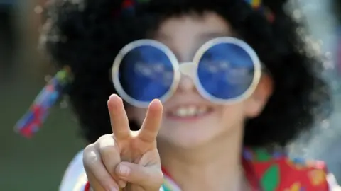 A boy in 1960s fancy dress with large circular glasses, headband and wig holds up a peace sign to camera.