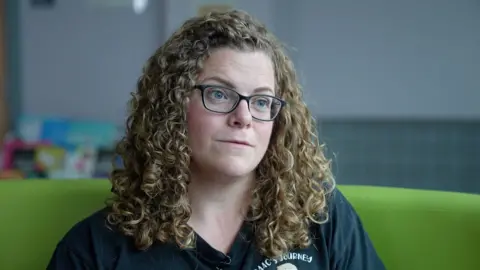 A woman with blonde curly hair wearing glasses and a black t-shirt which bears the logo Isaac's Journey. She is sitting on a green sofa, with a colourful stand containing leaflets behind her.