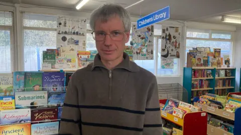 Jenny Kirk/BBC Mike Richmond stands in the library with shelves of children's books behind and around him.