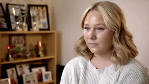 Headshot of Abby, who is pictured looking at a camera to the left hand side of the shot. She has shoulder-length, blonde, wavy hair and is wearing a cream ribbed jumper. She is pictured next to a wooden bookcase containing framed photographs and trophies.