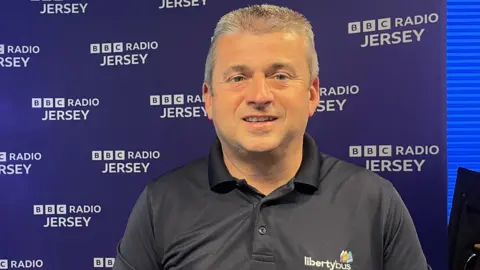 A mid shot of Kevin Hart, director of LibertyBus. He is smiling and wearing a polo shirt of the company. He is standing in front of a BBC Radio Jersey background.