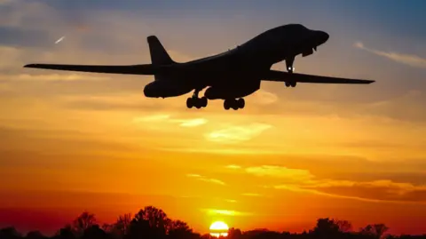The aeroplane is silhouetted in the sky against a stunning orangy-yellow sunset with the tops of a line of trees at the bottom of the picture and the sun disappearing on the horizon