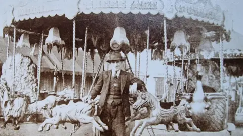 Jon Pullin A black and white of a man in a dark-coloured three-piece suit with a white shirt and dark-coloured tie. He is wearing a hat and is standing in front of a carousel leaning on two of the rides, one in the shape of a horse, the other a pig