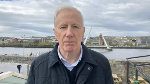 Shows a man in a dark jacket with a v-neck jumper and light blue shirt standing in front of the peace bridge in Derry