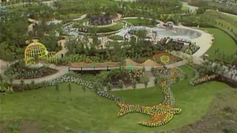 An aerial wide view of the National Garden Festival. There is a floral depiction of a bird in the bottom of the photo and behind it are various garden structures - including a bridge, water features and footpaths. 