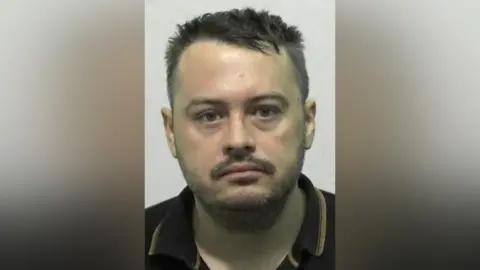 Mugshot of Aaron Keetley. He has scruffy short dark hair and a black moustache and beard,