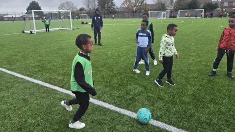 A group of young boys wearing jogging bottoms, trainers and jumpers are kicking a blue ball around a football pitch. One young boy wearing a green training bib looks about to kick the ball to his teammates. There are goalposts in the background and groups of people training.