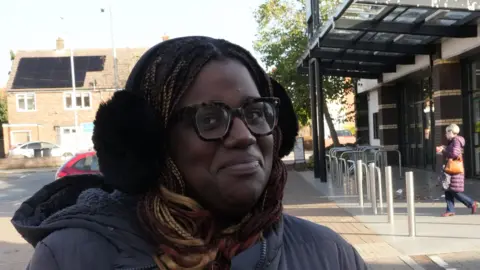 John Fairhall/BBC Tia Bell smiles at the camera as she stands outside a supermarket. She wears black ear muffs, glasses and wears a dark blue coat. Some shoppers can be seen leaving the building behind her. 