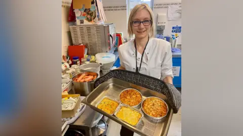 Hannah Hobbs-Chell Hannah Hobbs-Chell, who has blonde hair, black-rimmed glasses and a white shirt, is standing and smiling while holding a metal tray with oven gloves.