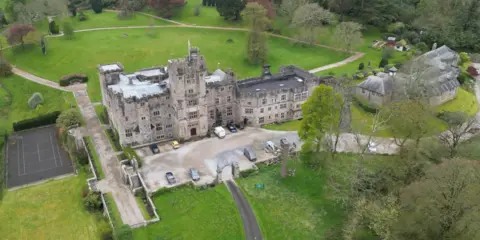 A grey stone, stately home stands on top of a hill. It has a crenelated turret with a union jack flying. To the right of the main house is a stable block. To the left there is a tennis court. It is surrounded by grass and a drive leads up to a gravel car park at the front of the property.
