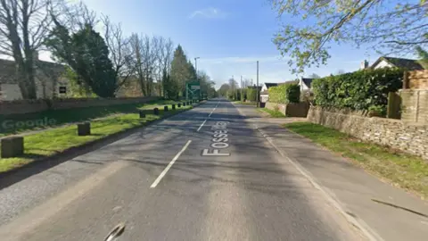 A road is pictured with no cars on it, and pavement and grassy verges either side.