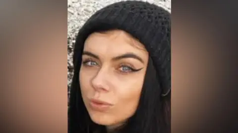 Holly Collinson looking at the camera wearing a black woolly hat. She has black hair.