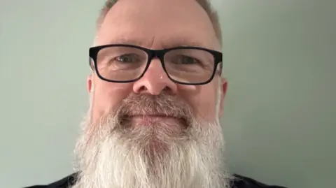 Royal Wolverhampton NHS Trust Chris has a long, white beard and is wearing glasses and smiling at the camera in a close-up selfie.