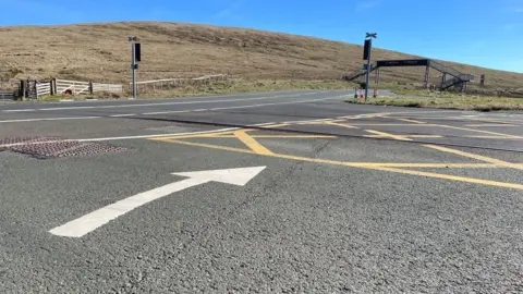 BBC A large white arrow on the road surface with the Bungalow tram lines behind, with the A18 Mountain Road in the Douglas direction. It is a sunny day.
