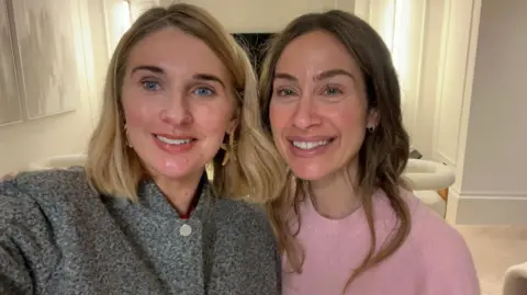 Two women smile into the camera, their heads together. the one on the left has shoulder length blonde hair and is wearing a dark grey top, the one on the right has long brown hair and is wearing a pale pink jumper.