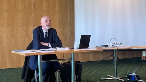 Andrew Turner/BBC John Dowsett, dressed in a dark blue suit, with white shirt and blue tie, sits at his desk. There is files of paper before him, and a laptop computer, and a projector, and a projector screen. The table has a wooden top and chrome legs.