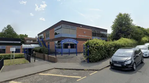 The entrance to Aston Fields Middle School is pictured from a street view. The entrance has a blue arch way with the name of the school written in white. The school building is predominately on two levels and made from red brick with blue windows.