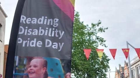 A sign reading "Reading Disability Pride Day". There is a girl smiling on the sign. To the left there is red and yellow bunting.