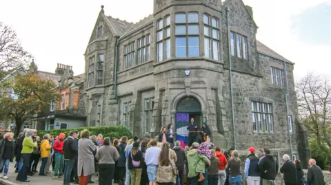 The Ladder An event opening of The Ladder. The Ladder is housed in a large old granite building and there is a man standing on its steps talking with a ribbon stretched across the entrance in front of him. A crowd is gathered, perhaps 40 people, and they are watching him. 