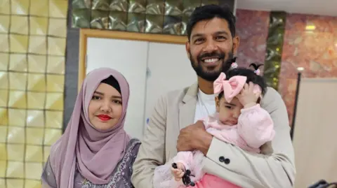 PA Media Muhammad Akhtar Hussain is smiling with his family. He is holding a small girl in his arms and his wife is stood next to him