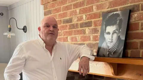 Peter Funnell A middle-aged man in a white shirt stands with his arm on a mantlepiece. On the mantlepiece there is a black and white photograph of a young man with dark hair, who is wearing a checked shirt and a jumper. 