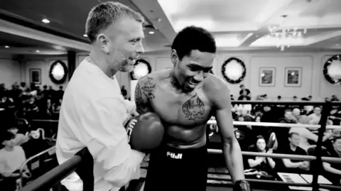 Clayton Chamberlain A black and white photo of a man supporting a young boxer in a ring, with a crowd watching.