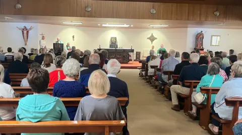 BBC Parishioners sitting in pews in the church as priests gather at the front to take the Mass. The alter can be seen in the picture alongside statues of Jesus and other figures, with crosses on the wall. 