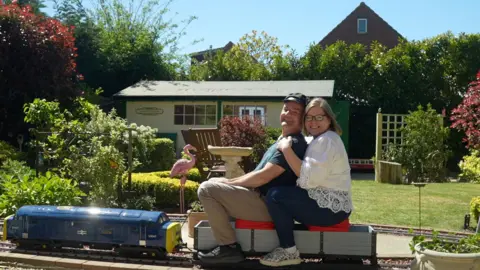 Jamie Niblock/BBC Angela, dressed in a white top and jeans, and Dave Walker, in brown-tan trousers and a T-shirt, sit on a miniature train carriage in their garden. There are bushes and a freshly cut green lawn. It is a sunny day.