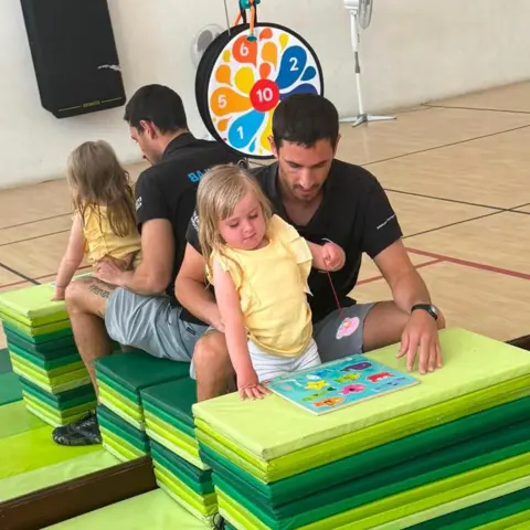 Warren Davies A young blonde girl wearing a yellow t short and white shorts. She is leant up against a stack of green play mats, playing with a coordination jigsaw game in front of her. She is being supported by a man with brown haor, wearing a black polo short and grey shorts. He is sat being her, on a another stack of green mats. Behind them both is a large mirrored wall.