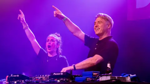 Day Fever  Vicky McClure and Jonny Owen with one arm aloft while standing behind DJ decks