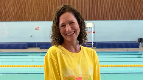 A woman with brown curly hair and a yellow tshirt stood in front of a swimming pool