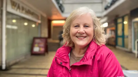Lesley smiles at the camera. She has shoulder-length blonde curly hair and is wearing a bright pink raincoat. Blurred behind her is a shopping centre she is sat in.
