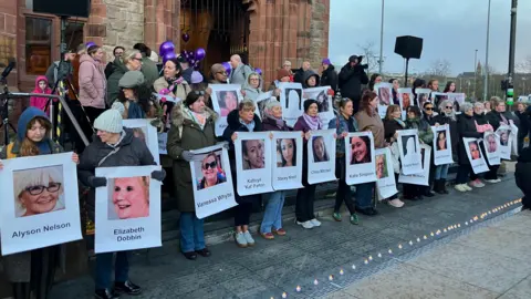 A group of women stand on the steps of Derry's Guildhall, each holding a poster of a woman killed in Northern Ireland