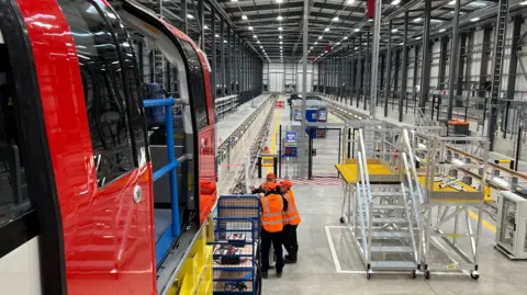 Inside a large warehouse-like train factory, with an underground train carriage on a raised platform to the left and work spaces to the right. Three workers in orange hi-vis jackets are standing in the centre of the work space.