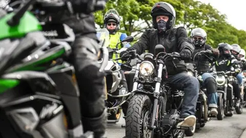 Riders on motorcycles line up next to each other, they sit on their machines wearing leather jackets and helmets.