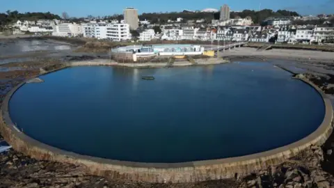 Tracey Cahill A nearly circular pool in bounded by a sea wall. It was photographed at low tide from the sea so a beach can be seen either side. There is also a white and turquoise building at the far end of the pool with steps leading down.
Blocks of flats, houses and other buildings in St Helier can be seen behind the pool.