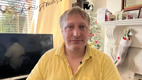 John Story sits in front of a TV and white fireplace in a living room decorated for Christmas. He has short fair hair and is wearing a yellow shirt. A Christmas tree and window can be seen in the background. A gold "Christmas" sign hangs near the window. A stocking hangs from the fireplace.
