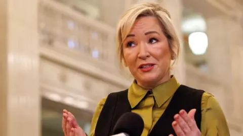 O'Neill is talking into a mic. She has blonde hair tied back and is wearing a green shirt and a black top over the top. 