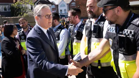 Reuters Keir Starmer shaking hands with one man wearing a high-vis jacket. The Home Secretary Shabana Mahmood is behind him also talking to other men,  wearing high-vis jackets. 