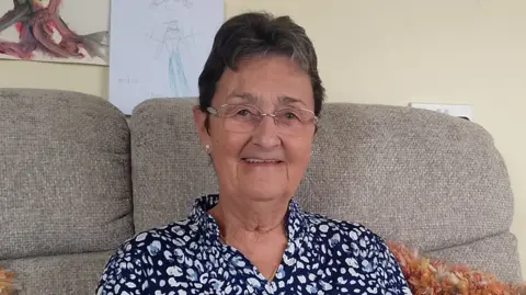 Woman with short black hair, wearing glasses and a navy and white patterned blouse. She is sitting on a cream sofa, an orange fluffy pillow can be seen over her left 