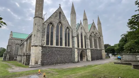 Andrew Turner/BBC A stone and flint church building with a green copper roof stands in a grassy churchyard.