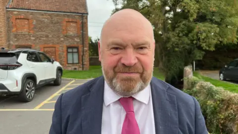 A man stood wearing a blue suit, pink shirt and pink tie looking at the camera. He has a neutral expression. Behind him is a white car and a house.