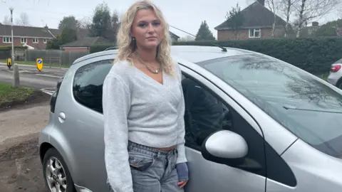 BBC Young woman with blonde hair wearing a grey v-neck jumper standing next to a grey Citroen C1 looking annoyed.