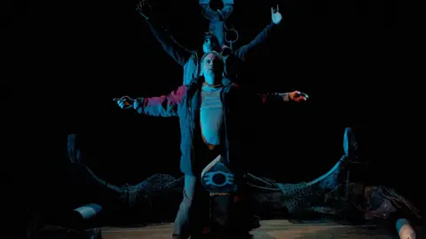 Two actors stand on stage dressed as fishermen, their arms outstretched, in front of a giant anchor.