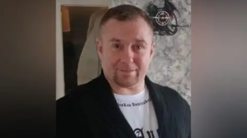 AS man with short brown hair and a short goatee beard wears a black jacket and white t-shirt.