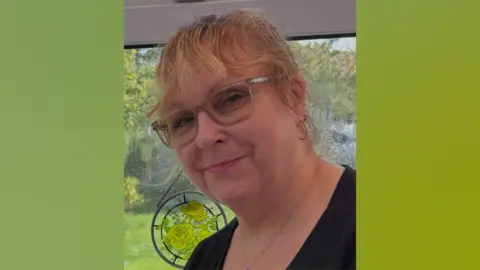 A woman with blonde hair. She has her hair tied back. She is wearing glasses and a black top. Behind her is a glass window and a suncatcher. 