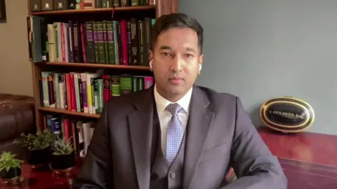 Ibraheem Dulmeer sits in an office in front of dark-wood furniture, including a bookcase full of hardback books. He has short dark hair and is wearing a smart, dark-grey three-piece suit, with a white shirt and blue tie. 