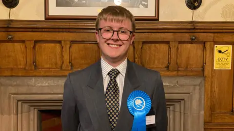 Marco Fawcett has light brown hair and glasses and is standing in front of a wooden panelled wall. He is wearing a suit and tie and he is smiling. He is also wearing a blue rosette.